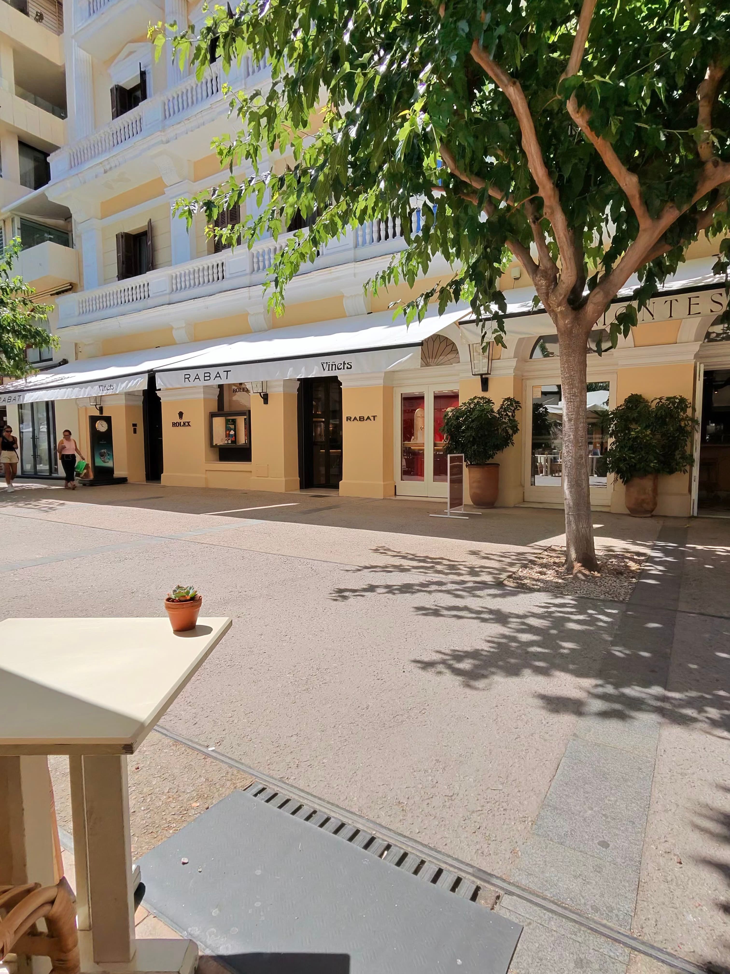 Sun-dappled pedestrian shopping street in Palma with Rabat jewellery and Rolex boutique under white awnings, shaded by a matu