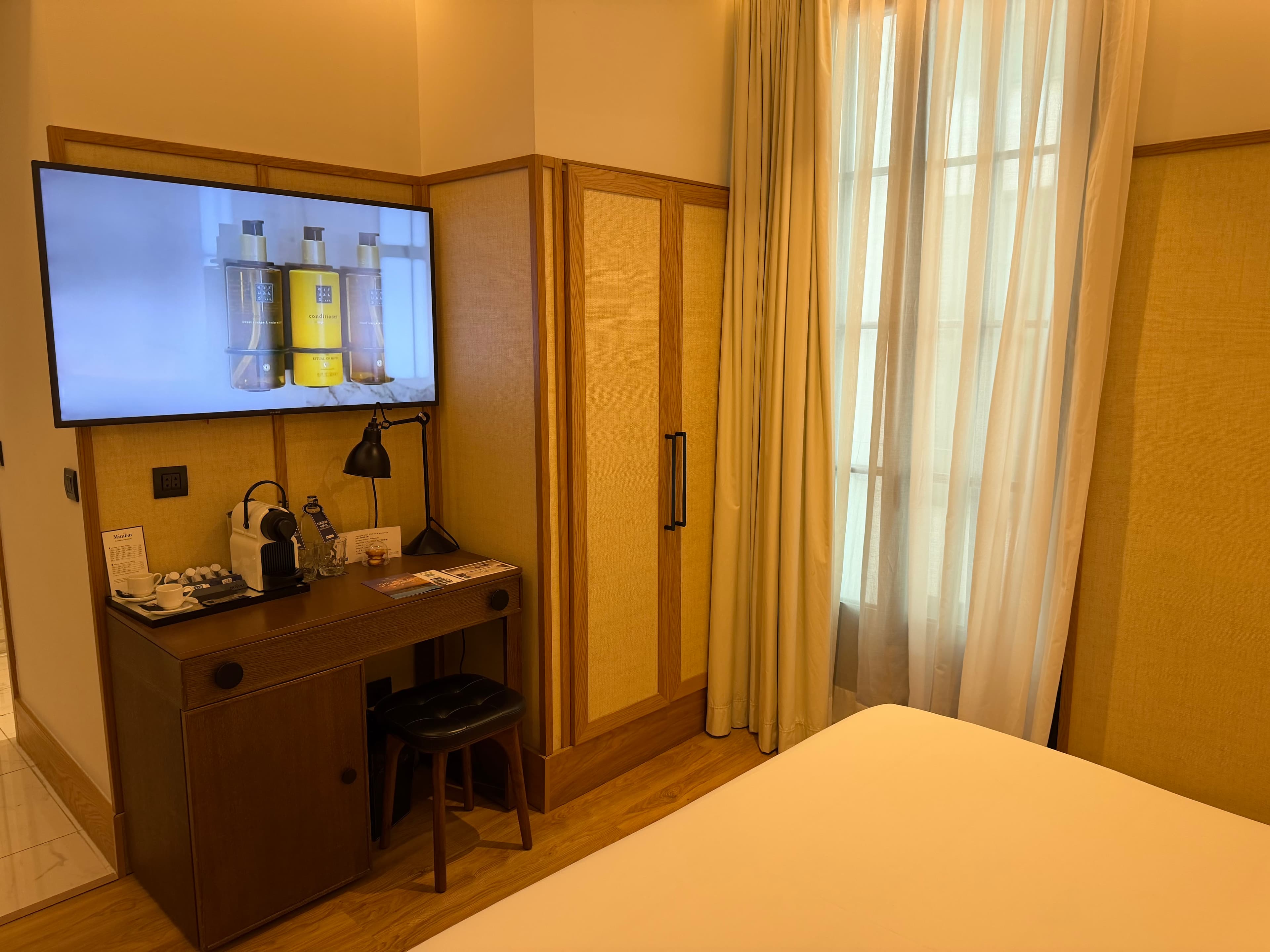 Boutique hotel room with warm wood tones, wall-mounted TV displaying amenities, Nespresso machine on desk, and sheer curtains