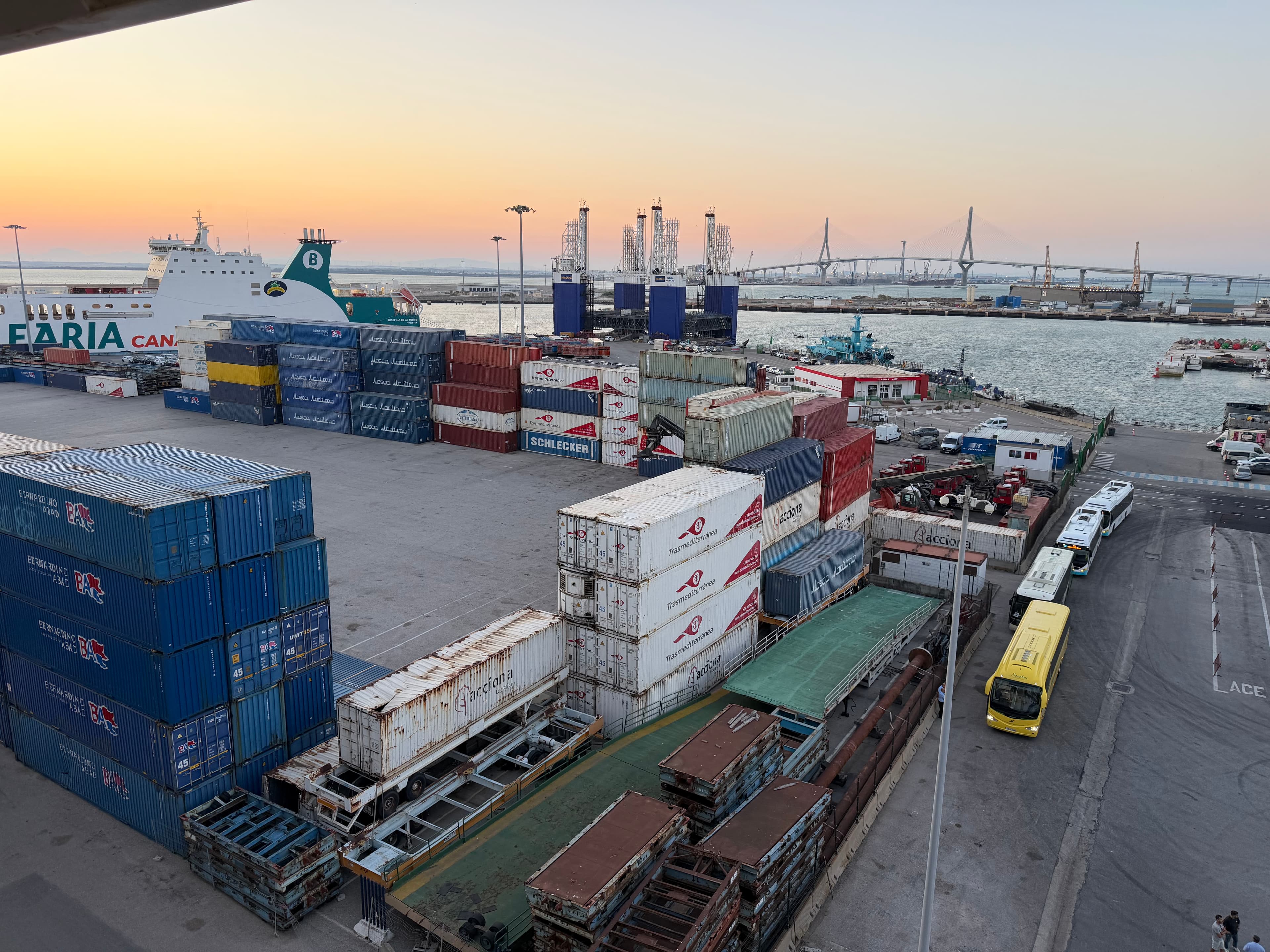 Busy Spanish port at dusk with stacked shipping containers, a Trasmediterránea ferry, buses, and a cable-stayed bridge glowin