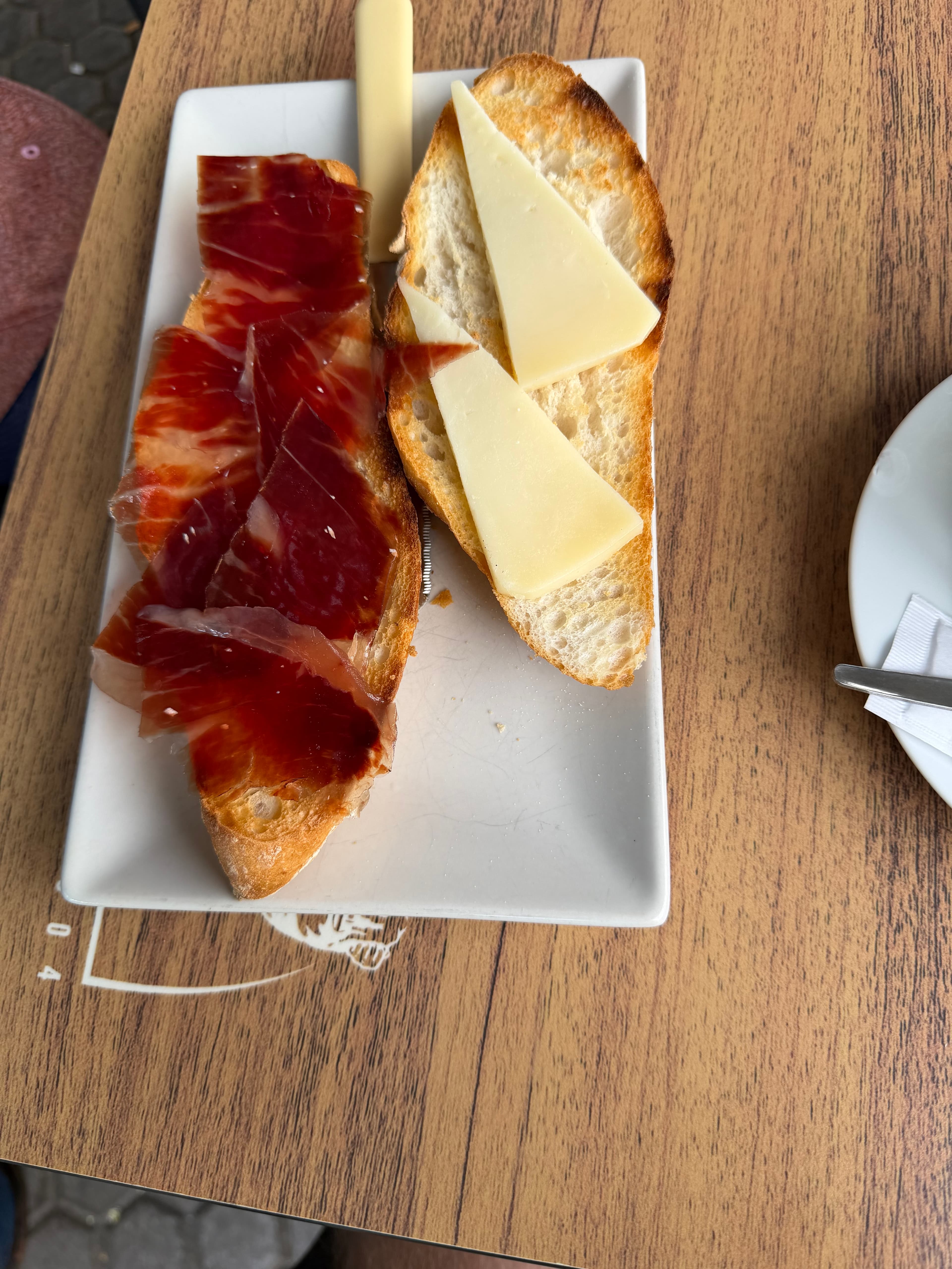 Two toasted baguette slices on a white rectangular plate — one topped with thin-sliced jamón ibérico, the other with wedges o