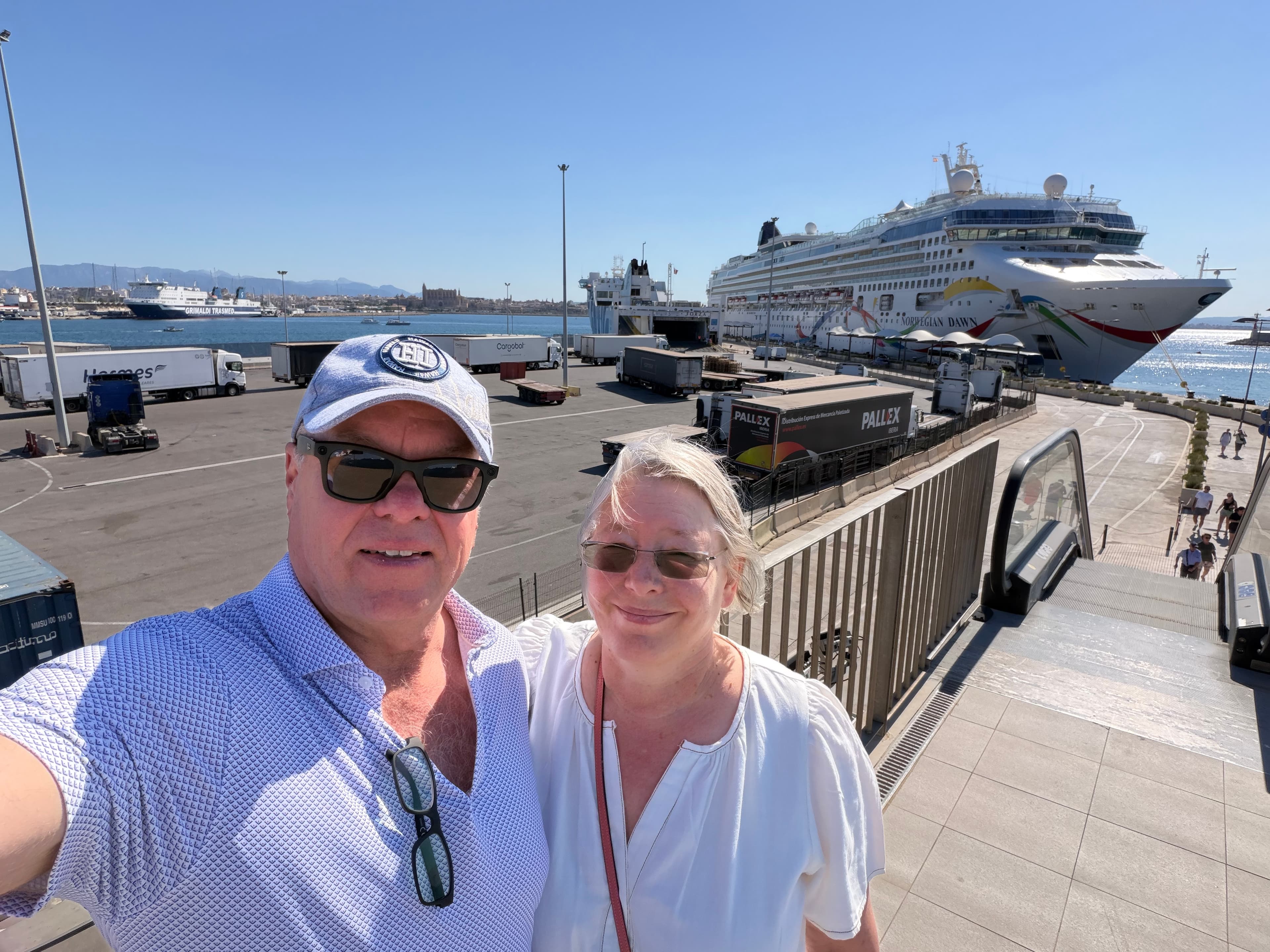 Smiling couple taking a selfie on a sunny cruise port terminal with the Norwegian Dawn cruise ship docked behind them under a
