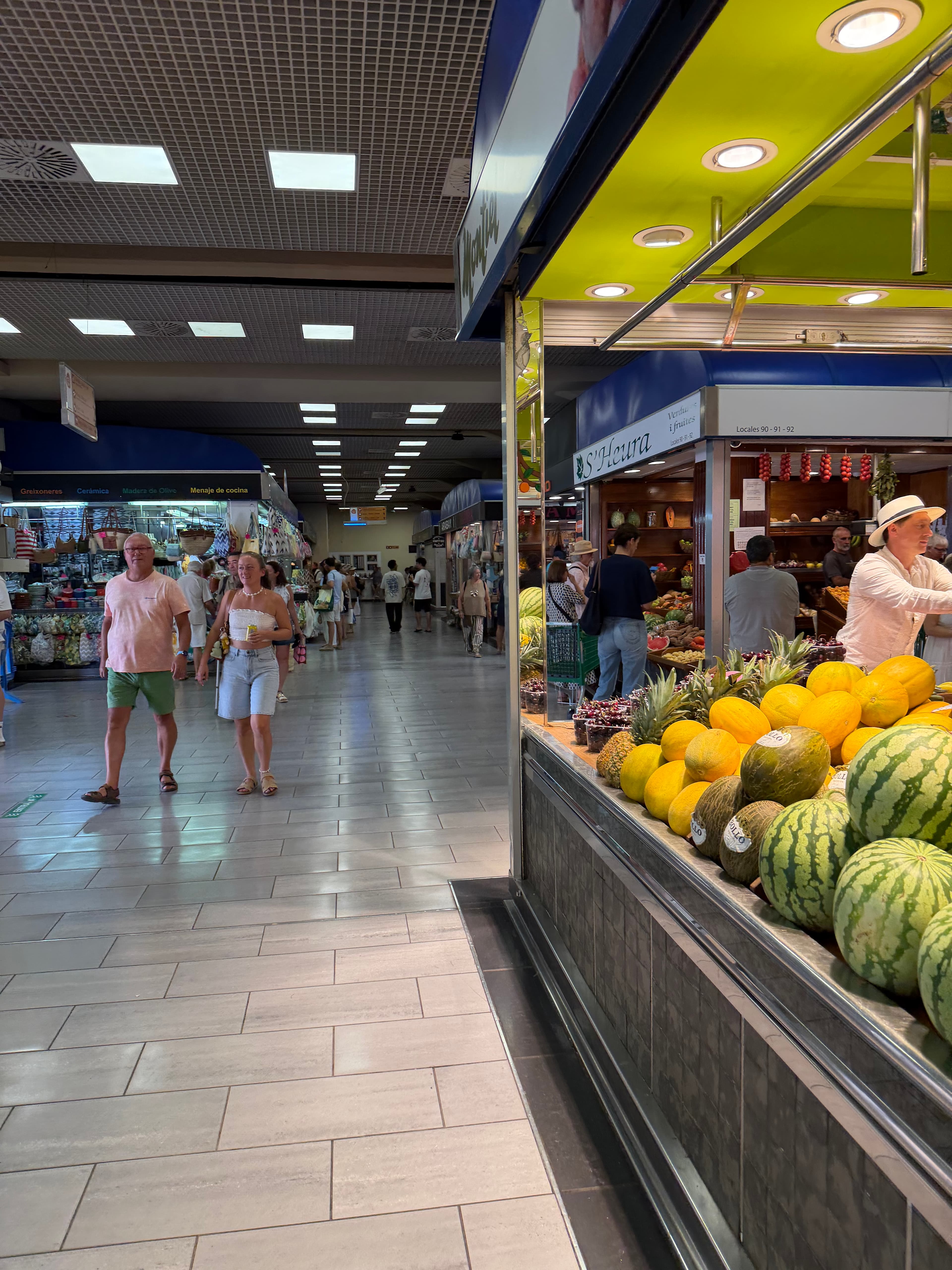 Busy indoor Spanish market hall with a fruit stall displaying watermelons and yellow melons in the foreground, shoppers strol