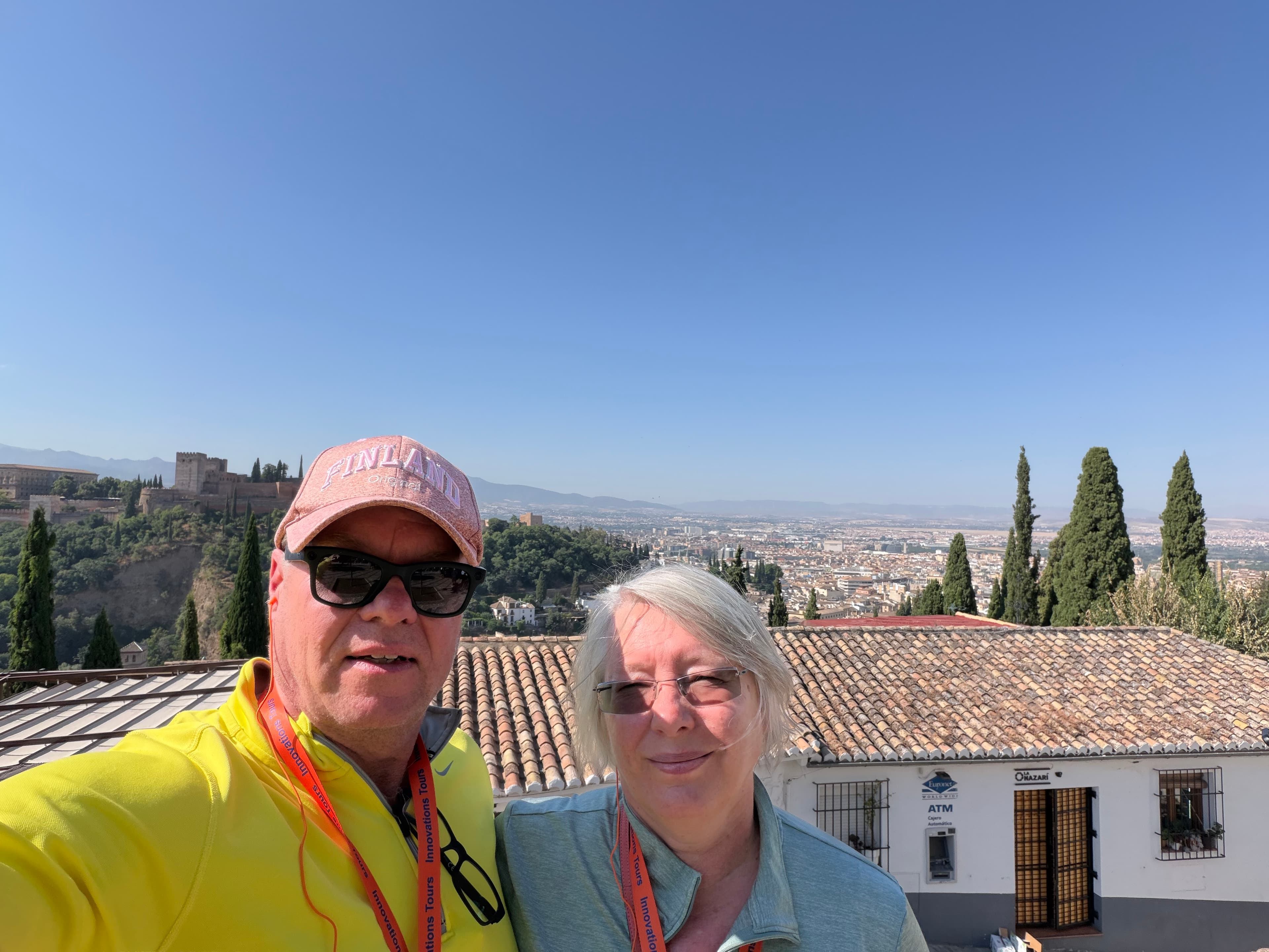 Couple wearing tour lanyards smiling in a sunny selfie with the Alhambra palace, cypress trees, and Granada cityscape behind