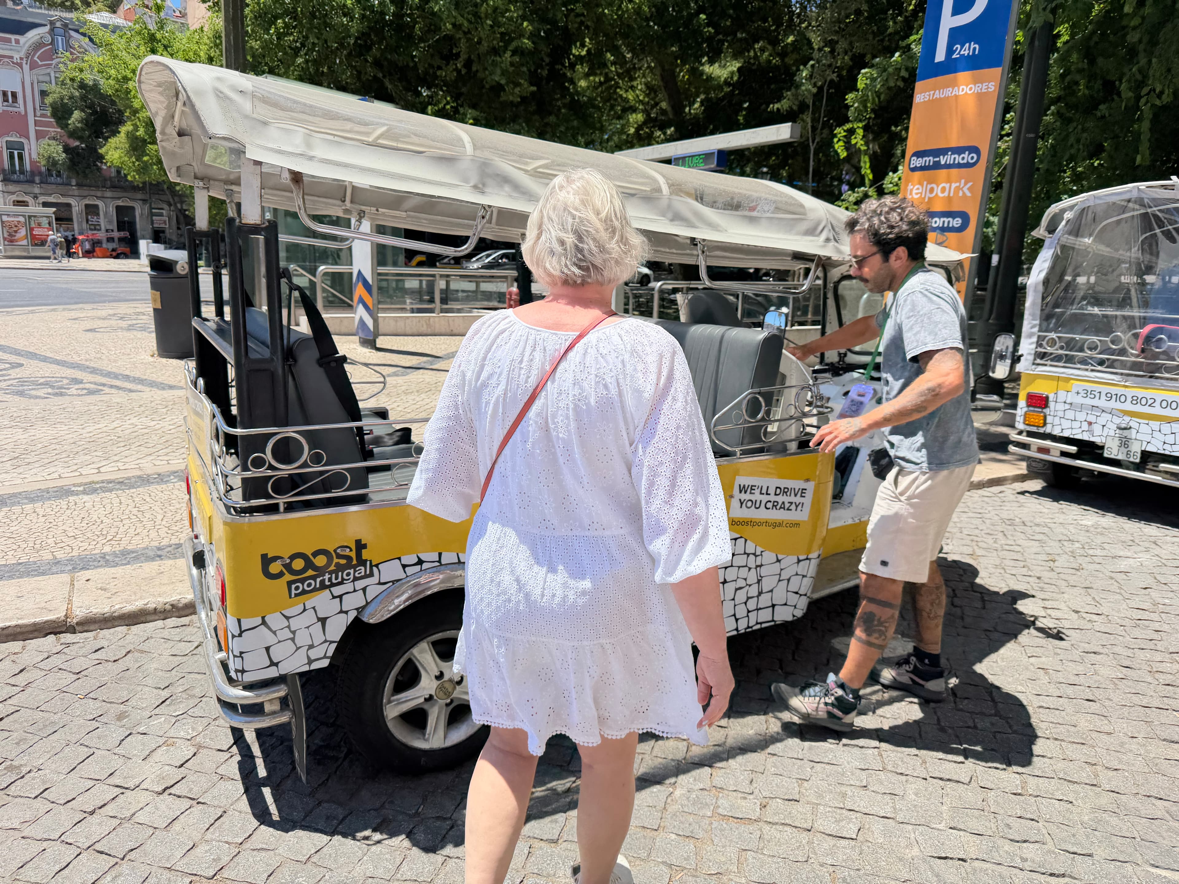 Woman in white dress approaching a yellow Boost Portugal tuk-tuk tour vehicle on a sunny Lisbon cobblestone street